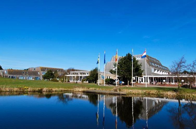View of Apartments STRANDHUYS and DE VUURTOREN in Hollum, Ameland, with serene pond and blue sky.